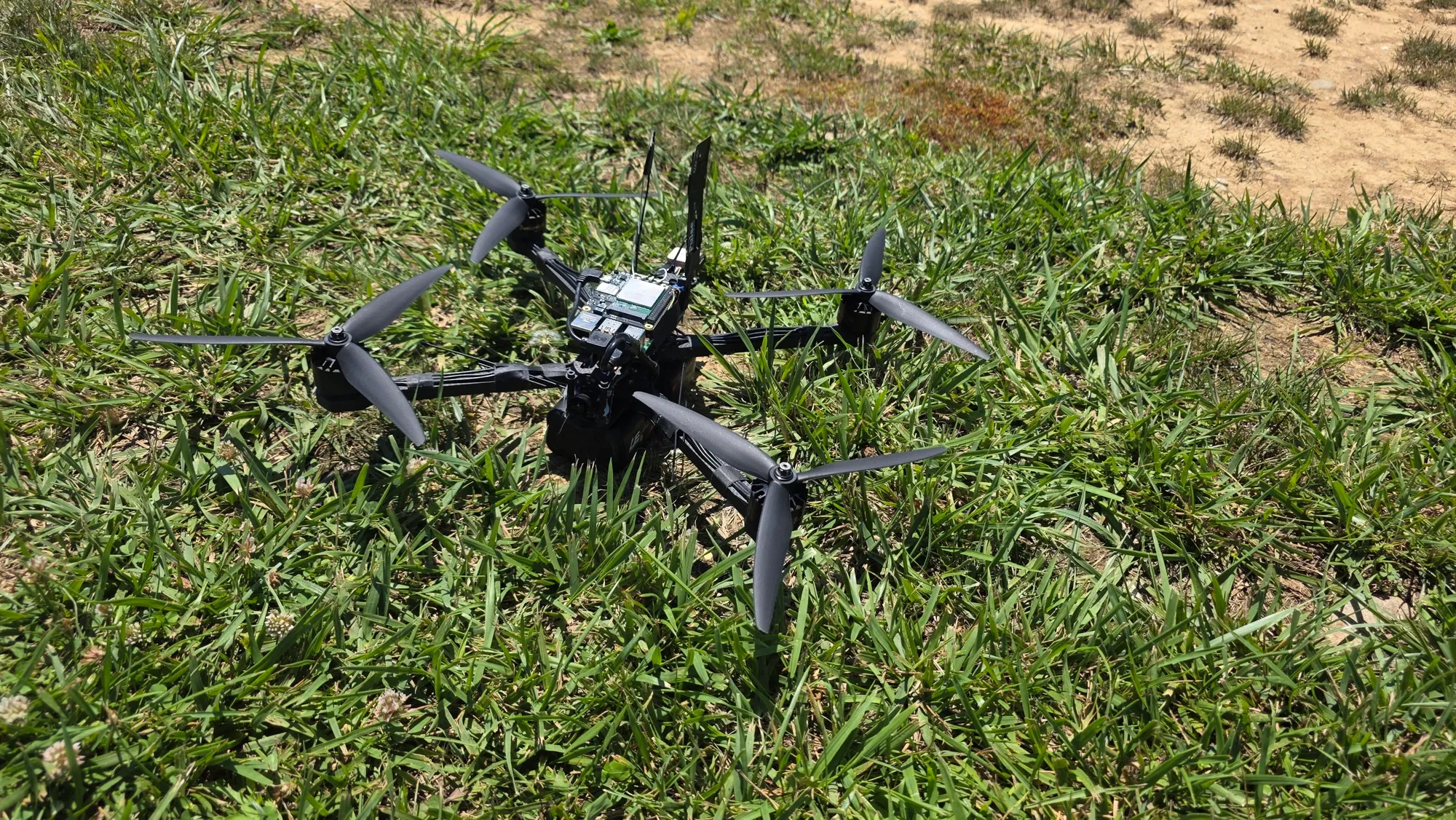 Low front view of a black quadcopter in grass, showing a small camera lens beneath stacked electronics boards.